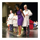 A new female student in a purple dress is white coated by a faculty member. 